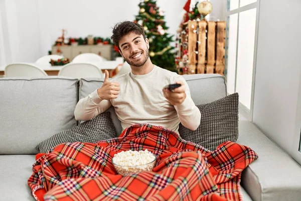 Young Hispanic Man Eating Popcorn Sitting Sofa Christmas Tree Smiling — Zdjęcie stockowe
