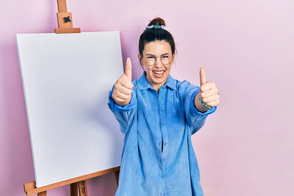 画家のイーゼルのそばに立つ若いヒスパニック系の女性は 手で積極的なジェスチャーを行うことを承認立って 親指を笑顔と成功のために幸せ 勝者のジェスチャー — ストック写真