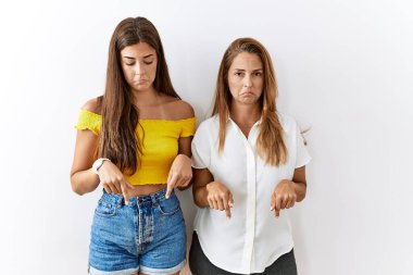 Mother and daughter together standing together over isolated background pointing down looking sad and upset, indicating direction with fingers, unhappy and depressed. 