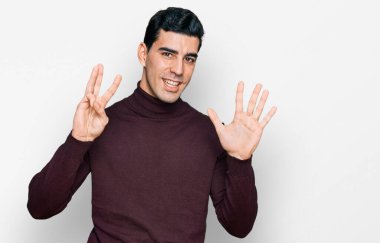 Yakışıklı, balıkçı yaka kazak giyen, kendini beğenmiş ve mutlu bir şekilde gülümseyen, sekiz numarayı gösteren İspanyol.. 