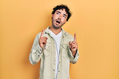Gündelik giysiler giyen genç İspanyol adam şaşırmış ve şaşırmış bir şekilde yukarı bakıyor ve parmaklarıyla işaret ediyor ve kollarını kaldırıyor.. 