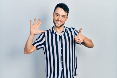 Latin asıllı genç adam gündelik giysiler giyip altı numaralı parmaklarıyla işaret ederken kendinden emin ve mutlu bir şekilde gülümsüyor.. 