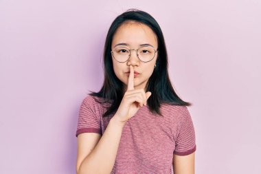 Gündelik kıyafetler ve gözlük takan genç Çinli kız parmakları dudaklarında sessiz olmak istiyor. sessizlik ve gizli konsept. 
