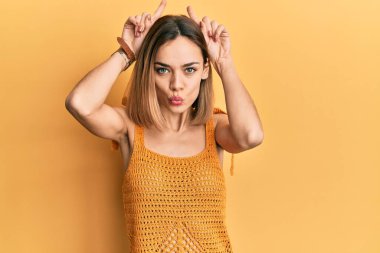 Kafkasyalı sarışın kadın, günlük sarı tişört giyiyor. Komik jestler yapıyor. Parmağını boynuz gibi kafasının üstünde gezdiriyor. 