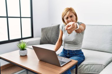 Orta yaşlı sarışın kadın evde dizüstü bilgisayar kullanıyor. Kameraya karşı kızgın ve sinirli. 