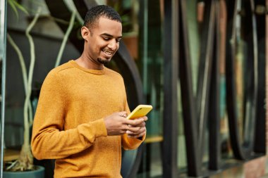 Genç Afrikalı Amerikalı adam şehirde akıllı telefon kullanarak mutlu bir şekilde gülümsüyor..