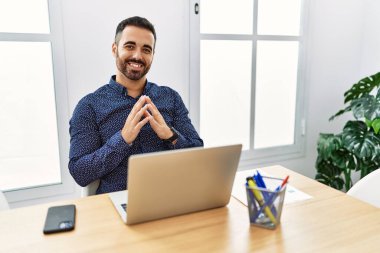 Ofiste sakallı, laptopla çalışan genç bir İspanyol adam. Elleri ve parmakları çapraz gülümseyerek rahat ve neşeli. Başarı ve iyimserlik 