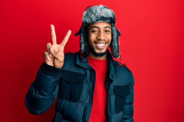 Sakallı, kışlık şapkalı ve paltolu genç Afrikalı Amerikalı iki numaralı parmaklarıyla işaret ederken kendinden emin ve mutlu bir şekilde gülümsüyor.. 
