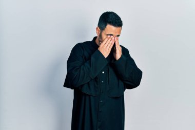 Sakallı yakışıklı, Katolik rahip cübbesi giyen, yorgunluk ve baş ağrısı için gözlerini ovuşturan, uykulu ve yorgun ifadeli bir İspanyol. görme sorunu 