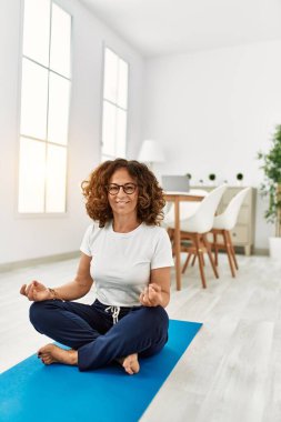 Orta yaşlı İspanyol kadın gülümseyerek evde yoga eğitimi alıyor.