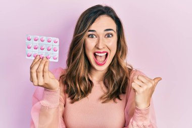 Genç İspanyol kız elinde haplar başparmağıyla yan tarafa bakıyor mutlu bir şekilde ağzı açık gülümsüyor. 