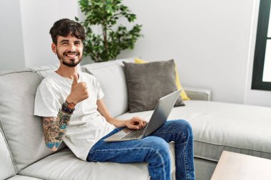 Kanepede oturan sakallı İspanyol adam elleriyle mutlu bir jest yapıyor. Kameraya bakıp başarıyı gösteren ifade. 