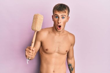 Young caucasian man shirtless holding shower sponge scared and amazed with open mouth for surprise, disbelief face 