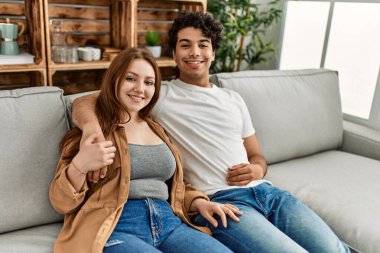 Mutlu gülümseyen ve sarılan genç çift evdeki kanepede oturuyor..