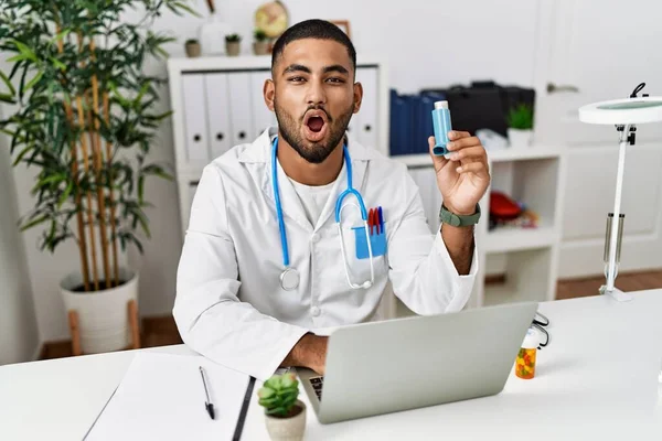 Young indian doctor holding medical asthma inhaler at the clinic scared and amazed with open mouth for surprise, disbelief face
