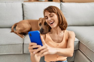 Kafkasyalı genç bir kadın akıllı telefonun yanında evde köpekle oturmuş selfie çekiyor.