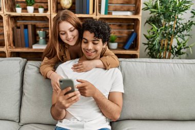 Mutlu gülümseyen ve sarılan genç çift evdeki kanepede oturuyor..