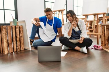 Resim yaparken gülümseyen iki öğrenci sanat okulunda yerde oturuyor..