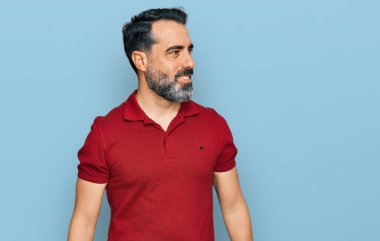 Orta yaşlı, sakallı, sıradan kırmızı tişörtlü, yüzünde gülümsemeyle yan yana bakan, doğal bir ifade. Kendinden emin gülen. 