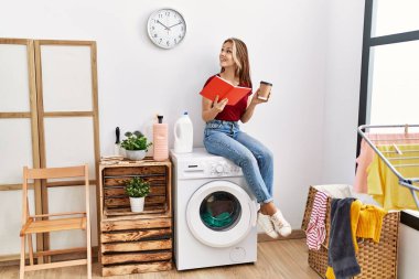 Young caucasian girl drinking coffee and reading book waiting for laundry sitting on whasing machine at home.