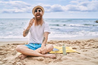 Genç kızıl adam dondurma yiyor kumsalda havlunun üzerinde oturuyor..