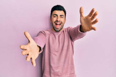 Gündelik giysiler giymiş genç İspanyol adam kameraya bakıp sarılmak için kollarını açmış gülümsüyor. Mutluluğu kucaklayan neşeli bir ifade.. 