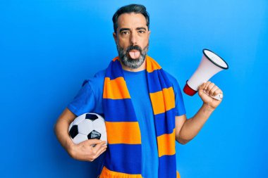 Orta yaşlı sakallı ve gri saçlı futbol holiganı topu tutuyor ve megafon kullanıyor. Komik bir ifadeyle dilini dışarı çıkarıyor.. 
