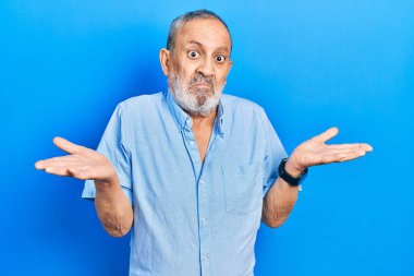 Sakallı, sıradan mavi tişörtlü, kafası karışmış, kolları ve elleri havada yakışıklı bir adam. şüphe kavramı. 