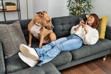 Beyaz bir kadın akıllı telefon kullanıyor. Kanepede köpeğiyle yatıyor.