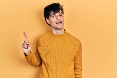 Yakışıklı hippi genç adam gündelik sarı süveter giyiyor, yüzünde kocaman bir gülümseme var, kameraya el ile işaret ediyor.. 