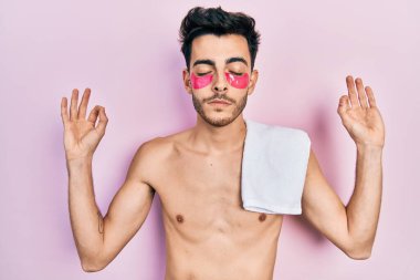 Havlu ve göz torbası takan genç İspanyol adam rahatla ve gülümse gözleri kapalı parmaklarıyla meditasyon hareketi yapıyor. yoga konsepti. 