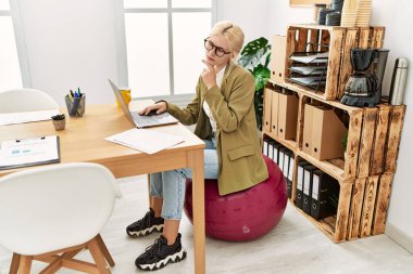 Ofiste çalışan beyaz, güzel bir iş kadını pilates topunun üzerinde oturuyor ciddi bir yüz ifadesi var eli çenesinde soru soran, kafa karıştırıcı bir fikir hakkında düşünceli. 
