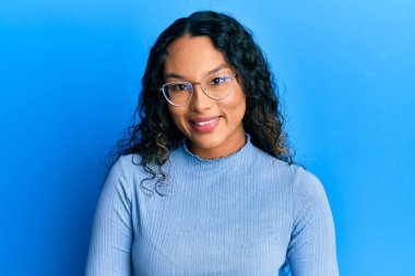 Yüzünde mutlu ve havalı bir gülümseme olan gündelik kıyafetler ve gözlük takan genç Latin bir kadın. şanslı kişi. 