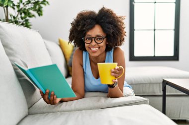 Genç Afrikalı Amerikalı kadın evde kahve içip kitap okuyor.