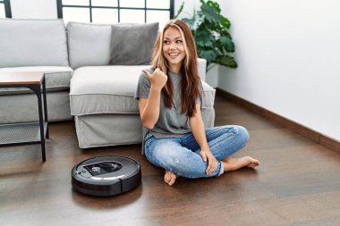 Young caucasian woman sitting at home by vacuum robot pointing thumb up to the side smiling happy with open mouth 