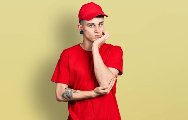 Kulakları genişlemiş, teslimat üniforması ve şapkası olan genç beyaz çocuk kollarını kavuşturarak depresyondan sıkılmış ve yorgun görünüyor.. 