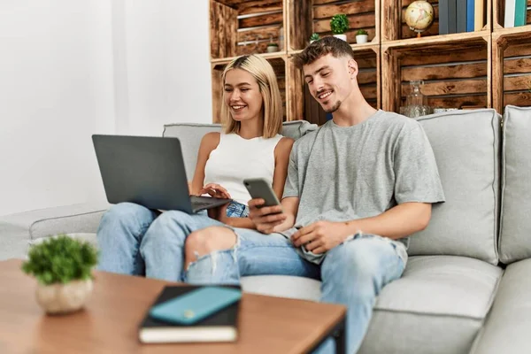 一对年轻的高加索夫妇坐在家里的沙发上 一边笑一边用笔记本电脑和智能手机开心地笑着 — 图库照片