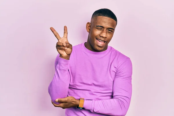 カジュアルなピンク色のセーターを着た若い黒人男性が カメラの前で手で勝利サインをする幸せそうな顔をして笑っている — ストック写真