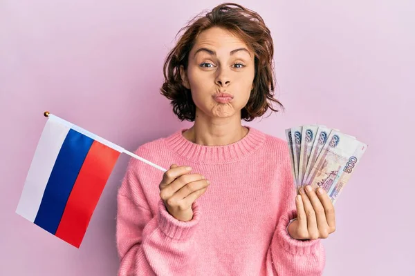 年轻的黑发女子举着俄罗斯国旗和卢布钞票 脸上带着滑稽的表情 鼓起双颊 嘴被空气吹胀 — 图库照片