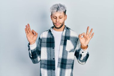 Modern boyalı saçlı, rahat gömlekli, gözleri kapalı gülümseyen ve parmaklarıyla meditasyon yapan genç İspanyol bir adam. yoga konsepti. 