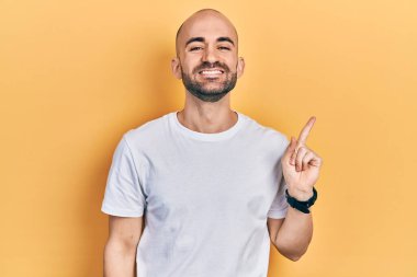 Yüzünde kocaman bir gülümseme olan günlük beyaz tişört giyen genç kel bir adam, kameraya el ile işaret ederek bakıyor.. 