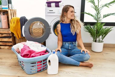 Çamaşır sepetinin yanında oturan genç ve güzel bir kadın yüzünde gülümsemeyle yan yana bakıyor, doğal bir ifade. Kendinden emin gülen. 