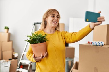 Orta yaşlı sarışın yeni evindeki akıllı telefonun yanında selfie çekiyor..