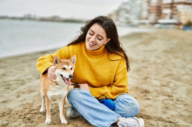 Güzel genç bir kadın sahilde mutlu Shiba Inu köpeğine sarılıyor.
