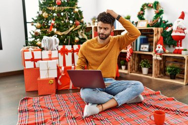 Arab young man using laptop sitting by christmas tree confuse and wondering about question. uncertain with doubt, thinking with hand on head. pensive concept. 