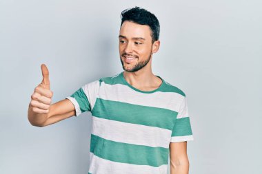 Latin asıllı, sıradan kıyafetler giyen, gururlu, gülümseyen, baş parmak kaldıran genç bir adam. 