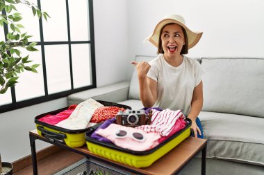 Güzel, orta yaşlı, İspanyol bir kadın valize yazlık giysiler koyuyor, gülümsüyor, gülümsüyor ve başparmağıyla yan tarafı işaret ediyor.. 