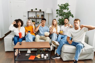 Group of people eating snack sitting on the sofa at home with angry face, negative sign showing dislike with thumbs down, rejection concept 