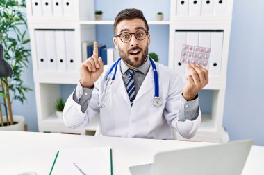 Handsome hispanic man wearing doctor uniform holding prescription pills surprised with an idea or question pointing finger with happy face, number one 