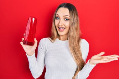 Young blonde girl holding shampoo bottle celebrating achievement with happy smile and winner expression with raised hand 